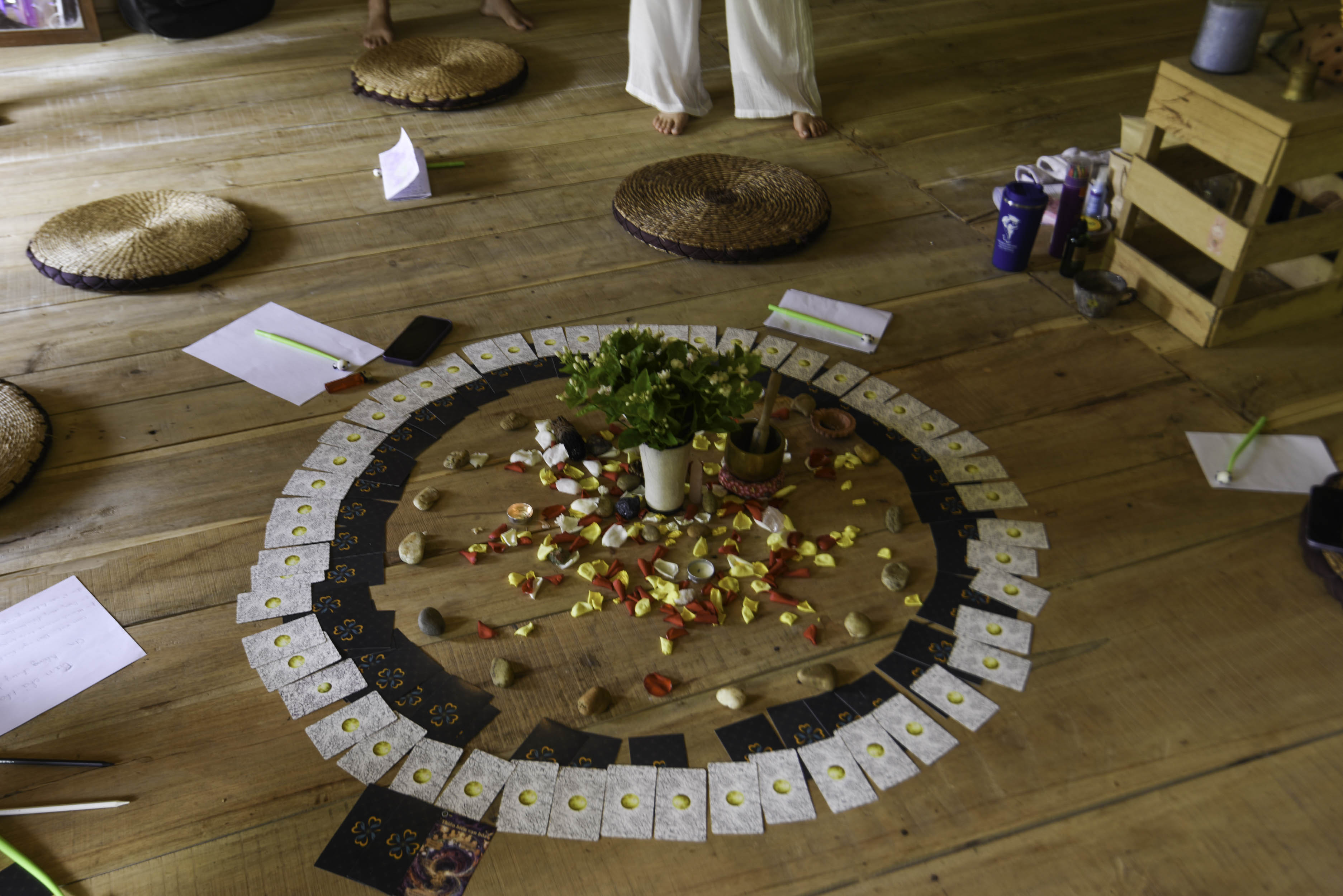 Cận cảnh mandala trên sàn gỗ: vòng thẻ bài, cánh hoa vàng đỏ, đá cuội, chậu hoa nhài, chuông xoay và giấy bút của người tham gia