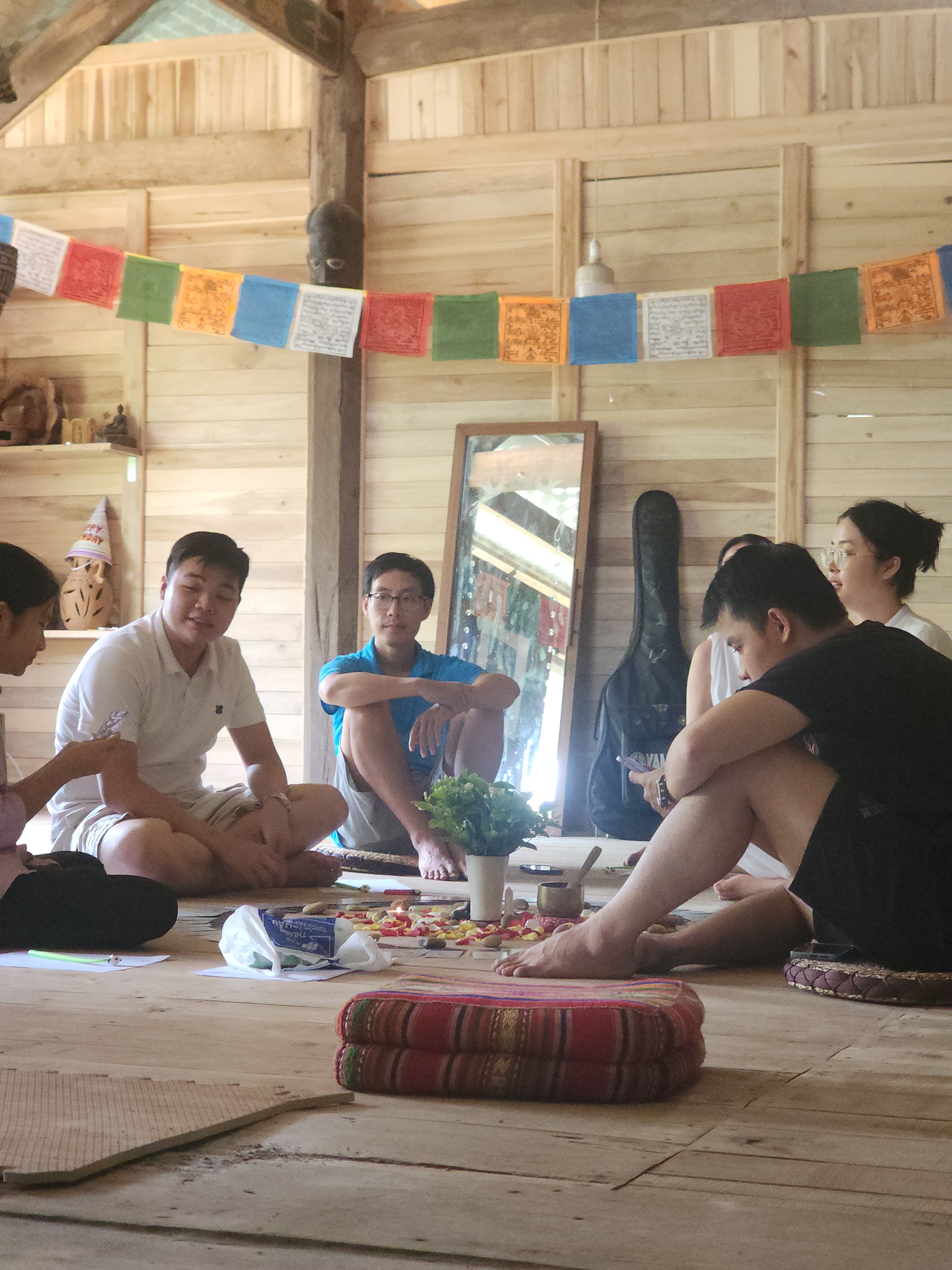 Vòng tròn ngồi bệt trong nhà sàn gỗ quanh mandala hoa và đá, có chuông xoay, gương và đàn guitar