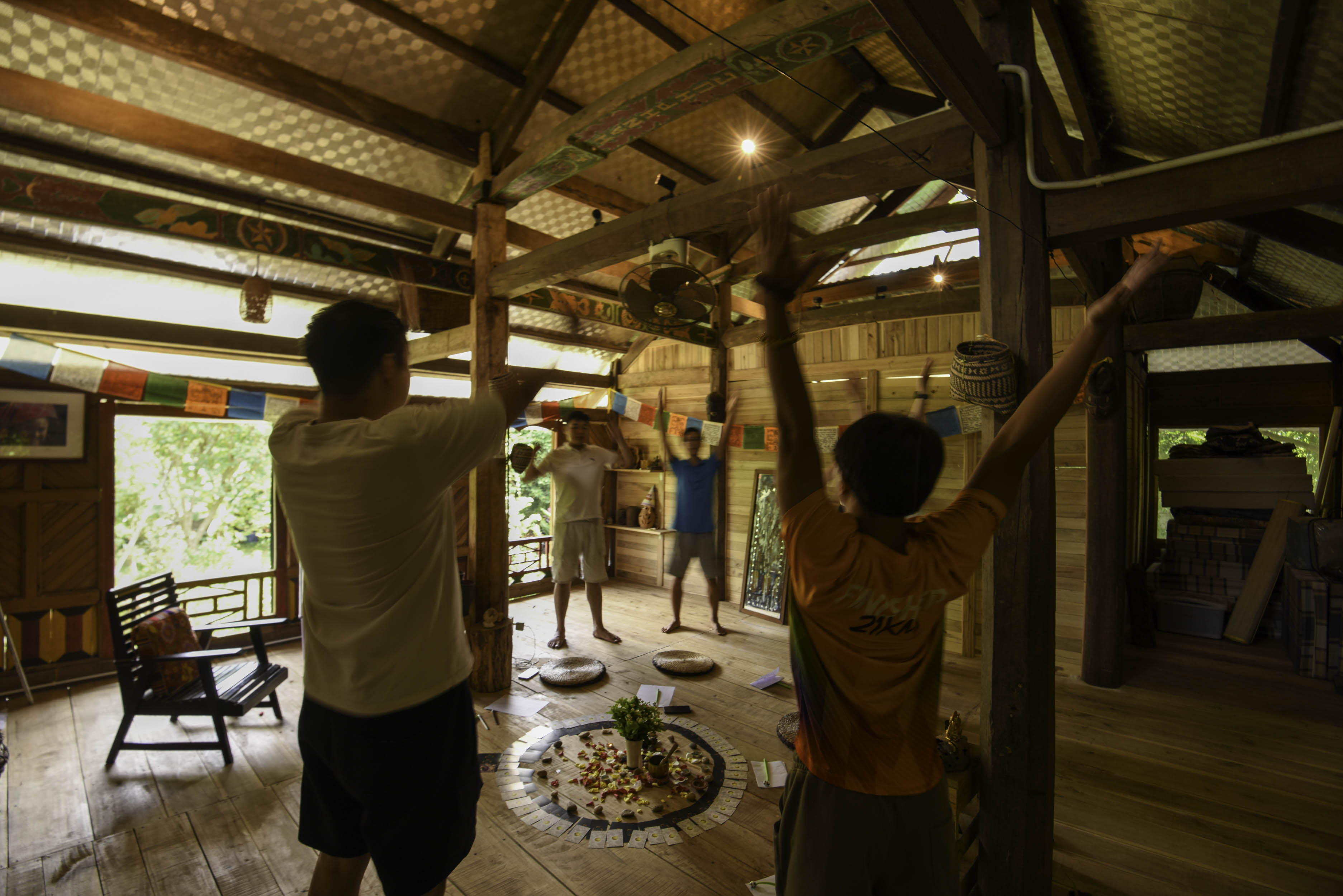 Mọi người đứng vươn hai tay lên cao trong nhà sàn gỗ, mandala trải dưới sàn và cờ Tây Tạng phía trên